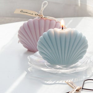 aesthetic minimal dreamy beach style aqua blue seashell shape soy pillar candle burning on holographic shell tray with lavender seashell candle in the back, and a black heart wick dipper on white round table