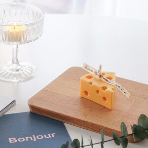 a slice of yellow cheddar cheese shape soy pillar candle with light yellow bluewine studio ribbon and eucalyptus leaf on Ikea mini wood cutting board, blue bonjour postcard, and a lit tealight candle in a glass coupe on white round table