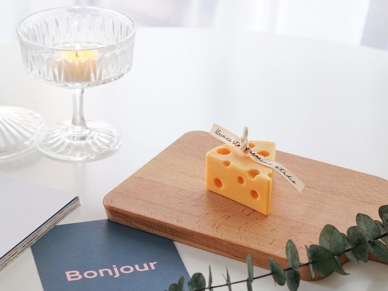 a slice of yellow cheddar cheese shape soy pillar candle with light yellow bluewine studio ribbon and eucalyptus leaf on Ikea mini wood cutting board, blue bonjour postcard, and a lit tealight candle in a glass coupe on white round table