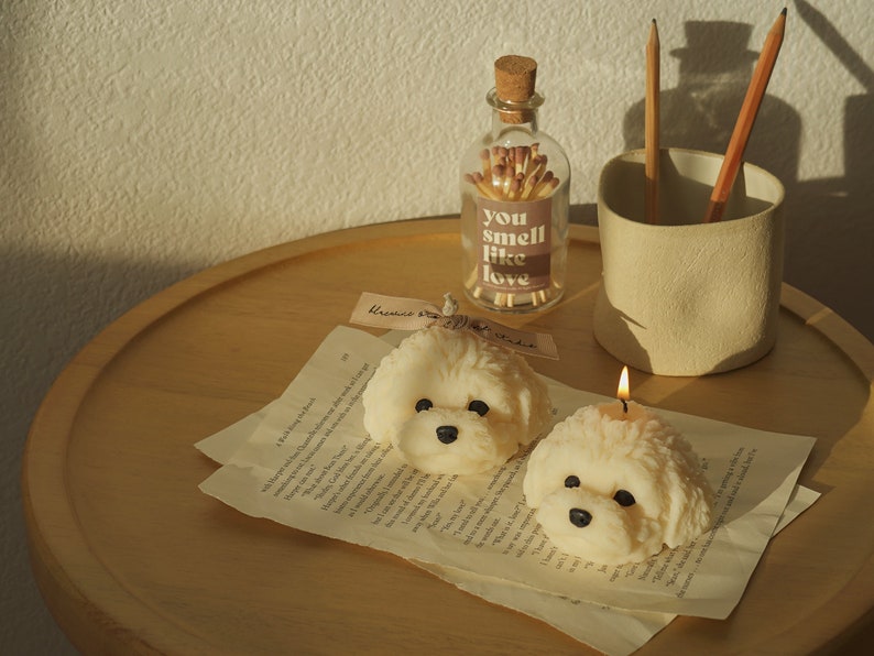 two white bichon candles placed on wooden table soy pillar candle with blue bluewine ribbon, a lit white bichon soy pillar candle in a blue mug, wood color pencils, and two house lamps on the white round table