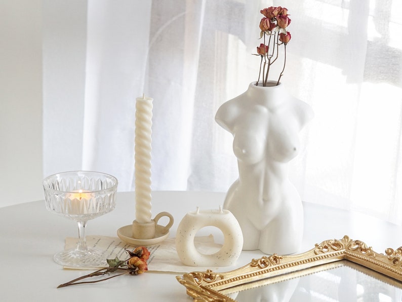 white twisted swirl taper candle in a ceramic candle holder, orange rose dried flowers, ring shape dotted candle, a lit tealight in a coupe glass, rectangular gold french mirror tray, a feminine body shape white ceramic vase on white round table