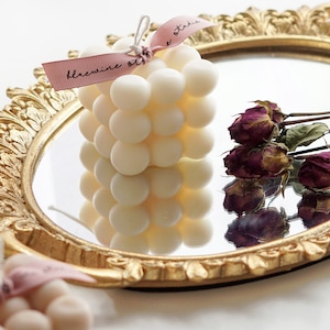 There is a aesthetic french style vanity mirror on the bottom and there is a handmade cute square bubble cube soy wax candle with bluewine studio ribbon and dried flowers on top of the white display table.