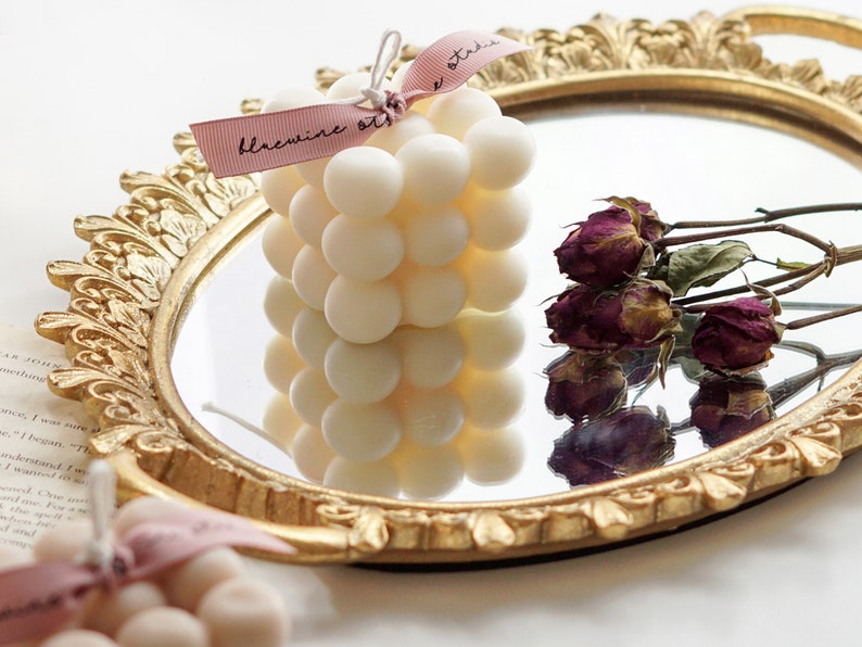 There is a aesthetic french style vanity mirror on the bottom and there is a handmade cute square bubble cube soy wax candle with bluewine studio ribbon and dried flowers on top of the white display table.