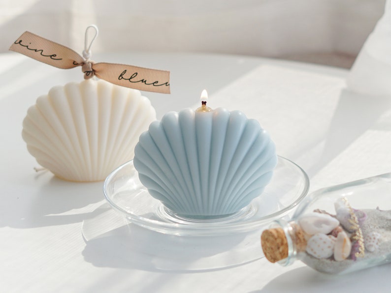 aesthetic minimal dreamy beach style mood from a lit aqua blue seashell shape soy pillar candle on a clear dish, and a white seashell candle with beige bluewine studio ribbon