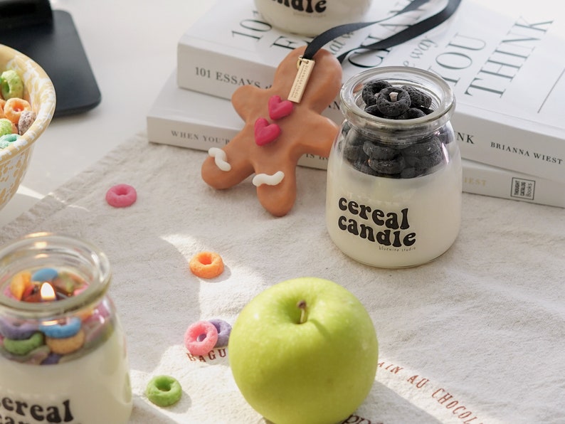 a lit colorful cereal candle, fruit loops wax melts, a green apple, oreo candle, gingerbread scented wax, 101 Essays that will change the way you think by Brianna Wiest placed on kitchen cotton fabric