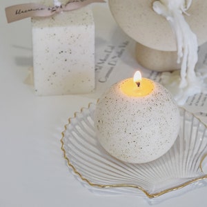a lit dotted white sphere ceramic pattern soy pillar candle on a gold rim clear seashell tray and a dotted white square pillar candle with beige bluewine studio ribbon