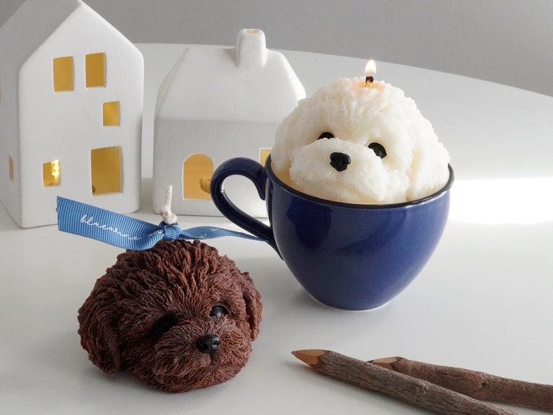 chocolate poodle soy pillar candle with blue bluewine ribbon, a lit white bichon soy pillar candle in a blue mug, wood color pencils, and two house lamps on the white round table