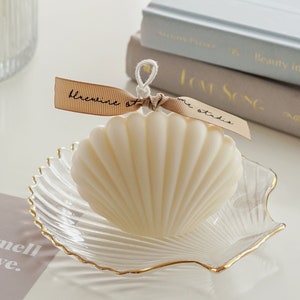 dreamy aesthetic minimal white seashell soy pillar candle on a gold rim clear shell tray, two books, you smell like love beige postcard