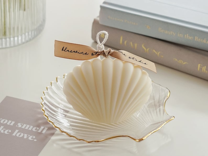 dreamy aesthetic minimal white seashell soy pillar candle on a gold rim clear shell tray, two books, you smell like love beige postcard