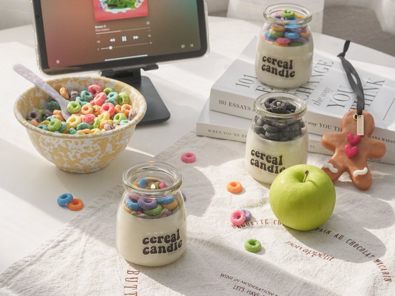 a lit colorful cereal candle, fruit loops wax melts, a green apple, oreo candle, gingerbread scented wax placed on kitchen cotton fabric, yellow crow canyon salad bowl filled with froot loops, and ipad mini.