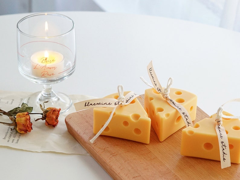 a three yellow orange color of cute cheese shaped design handmade candles with bluewine studio text written on a ribbon, red color dried flowers, old paper sheets and tealight candle in a wine glass  on the white aesthetic minimal simple table desk.