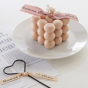 There is neutral tone beige color dreamy aesthetic minimal style square bubble cube handmade soy wax candle with bluewine studio ribbon with heart shaped candle wick dipper displayed on the table.