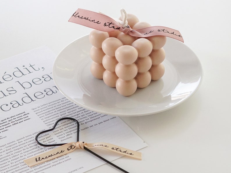 There is neutral tone beige color dreamy aesthetic minimal style square bubble cube handmade soy wax candle with bluewine studio ribbon with heart shaped candle wick dipper displayed on the table.