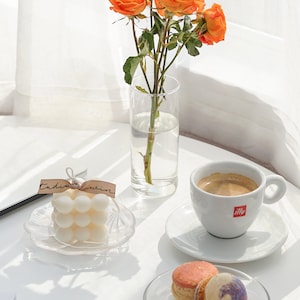 beautiful sunlight reflecting on the white round ikea minimal table with variety of display objects like orange roses flowers, handmade soy wax bubble cube candle, sweet macaron desserts and coffee on the table.