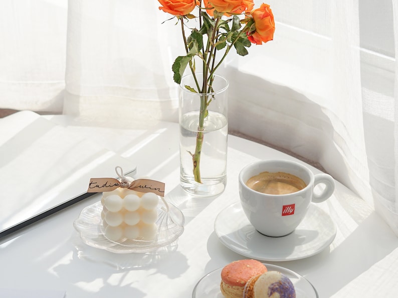 beautiful sunlight reflecting on the white round ikea minimal table with variety of display objects like orange roses flowers, handmade soy wax bubble cube candle, sweet macaron desserts and coffee on the table.