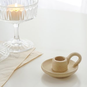 a dotted neutral beige color ceramic taper candle holder and a lit tealight candle in a coupe glass placed on book pages on white round table