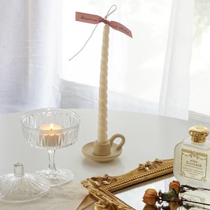 white twisted soy pillar candle in a dotted beige ceramic taper candle holder, a lit tealight candle in a coupe glass, and yellow rose dried flowers and santa maria novella perfume placed on rectangular gold french mirror tray on white table