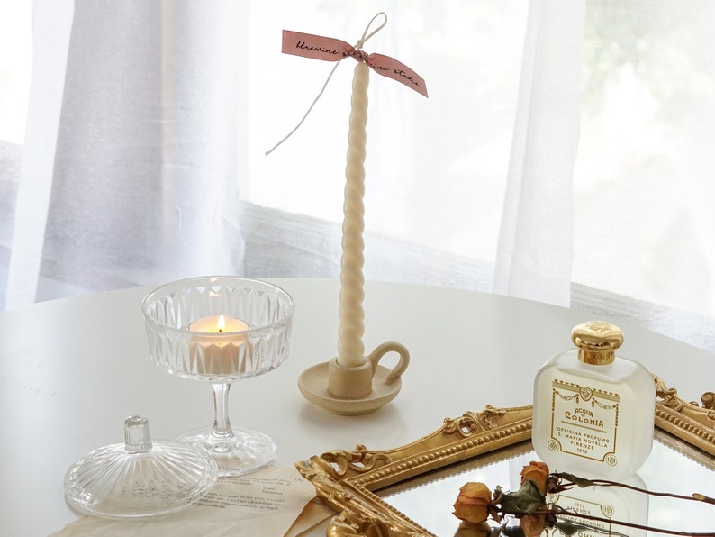 white twisted soy pillar candle in a dotted beige ceramic taper candle holder, a lit tealight candle in a coupe glass, and yellow rose dried flowers and santa maria novella perfume placed on rectangular gold french mirror tray on white table