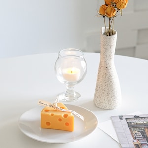 a slice of yellow cheddar cheese shape soy pillar candle on a white ceramic plate, a lit tealight candle in a mini glass, a postcard, yellow dried roses in a ceramic rough texture vase on white round table