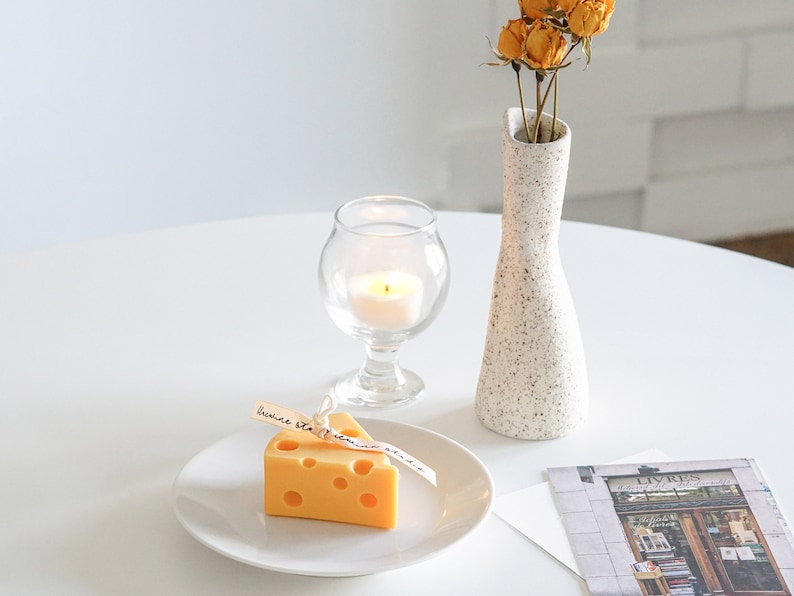 a slice of yellow cheddar cheese shape soy pillar candle on a white ceramic plate, a lit tealight candle in a mini glass, a postcard, yellow dried roses in a ceramic rough texture vase on white round table