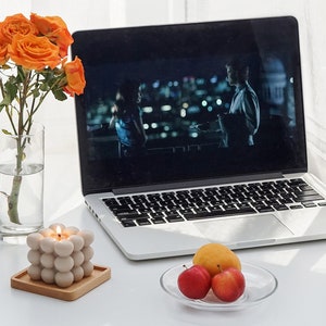 There are orange apricot color flowers in a vase, beige mono color square bubble cube candle, peaches and apple laptop on the desk.