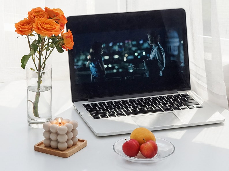 There are orange apricot color flowers in a vase, beige mono color square bubble cube candle, peaches and apple laptop on the desk.