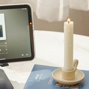 a lit white soy pillar taper candle in a beige neutral color candle holder placed on blue book and ipad mini playing the song Japanese Denim by Daniel Ceaser