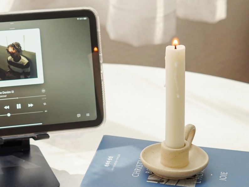 a lit white soy pillar taper candle in a beige neutral color candle holder placed on blue book and ipad mini playing the song Japanese Denim by Daniel Ceaser