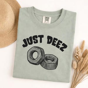 May include: Sage green t-shirt with the text "JUST DEEZ" in black, above a graphic of two nuts. The shirt has a crew neck and a tag that says "COMFORT COLORS". A straw hat and dried plants are in the background.
