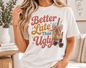 May include: A white short-sleeve t-shirt featuring the text Better Late Than Ugly in red and gold script. The graphic includes an iced coffee with a leopard print straw and pink bow, a red lipstick tube, and heart-shaped sunglasses.