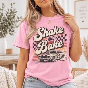 Puede incluir: Una camiseta de manga corta de color rosa claro con un estampado gráfico que incluye el texto Shake and Bake, un patrón de cuadros y un coche de carreras de estilo vintage con el número 26 en el lateral, sobre un fondo inspirado en el atardecer.