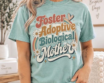 May include: A teal short-sleeve t-shirt featuring a graphic print with the text Foster Adoptive Biological Mother. The design includes a rainbow, white daisies, and small stars in a retro font style with muted red, orange, and blue colors.