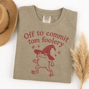以下が含まれることがあります： 赤色の文字で「Off to commit tom foolery」と書かれたセージグリーンのTシャツ。テキストの下には、魔女の帽子をかぶった漫画のカエルがプリントされています。このシャツは柔らかい素材で作られています。