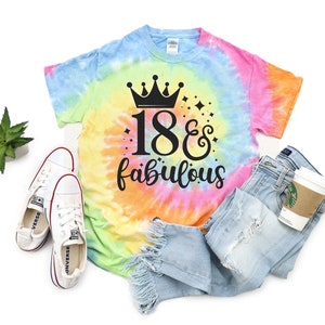 Puede incluir: Una camiseta tie-dye con una corona y el texto "18 & fabulous". La camiseta se muestra sobre un fondo blanco con un par de zapatillas blancas y un par de pantalones vaqueros azules.