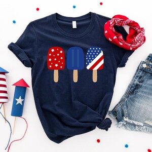 May include: A navy blue t-shirt with a graphic of three popsicles. The popsicles are red, white, and blue and have a star pattern.