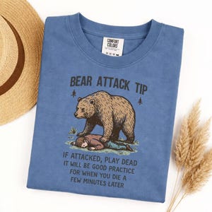 May include: A blue t-shirt with the text "BEAR ATTACK TIP" and a graphic of a bear and a person. The text below reads "IF ATTACKED, PLAY DEAD IT WILL BE GOOD PRACTICE FOR WHEN YOU DIE A FEW MINUTES LATER."