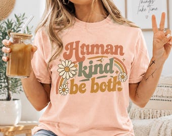 May include: A peach-colored short-sleeve t-shirt featuring the text "Human kind be both" in a retro font. The graphic includes a multi-colored rainbow, small star accents, and three white daisy flowers with yellow centers printed on the front.