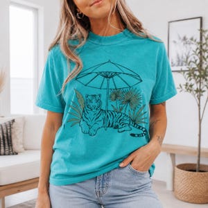 May include: A turquoise short-sleeve t-shirt featuring a black line art graphic of a tiger resting under a beach umbrella surrounded by palm leaves. The shirt has a crew neckline and a relaxed fit, showcasing a detailed illustration on the front.