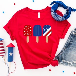 May include: Red t-shirt with a graphic of three popsicles. The popsicles are decorated with red, white, and blue stars and stripes.