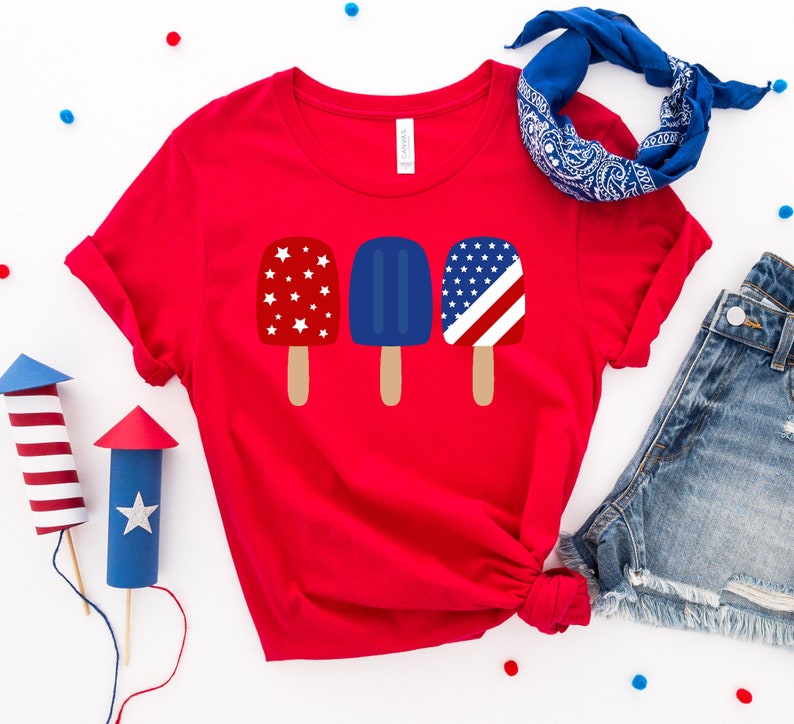 May include: Red t-shirt with a graphic of three popsicles. The popsicles are decorated with red, white, and blue stars and stripes.