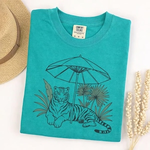 May include: Teal Comfort Colors t-shirt featuring a tiger resting under an umbrella, with palm fronds and other foliage. The graphic is in a dark green color. A straw hat is partially visible in the upper left corner.