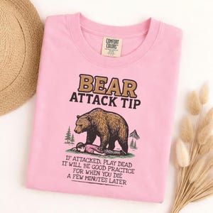 May include: Pink Comfort Colors t-shirt with a humorous graphic. The graphic reads "BEAR ATTACK TIP" above an illustration of a bear and a person. Below the illustration is the text "IF ATTACKED, PLAY DEAD..."