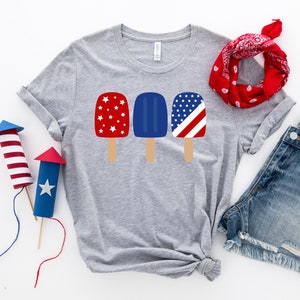 May include: A gray t-shirt with a design of three popsicles. The popsicles are red, white, and blue with stars and stripes. The shirt is laid out on a white surface with a red bandana and denim shorts.