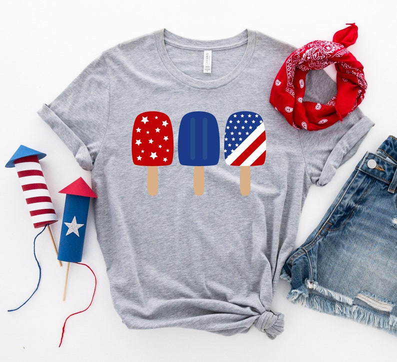 May include: A gray t-shirt with a design of three popsicles. The popsicles are red, white, and blue with stars and stripes. The shirt is laid out on a white surface with a red bandana and denim shorts.