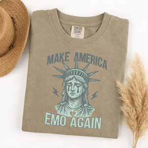 May include: A Comfort Colors olive green t-shirt featuring the text "MAKE AMERICA EMO AGAIN" above a distressed illustration of the Statue of Liberty with tears and a broken heart. The shirt has a crew neck and short sleeves.