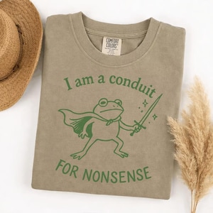 May include: A Comfort Colors olive green t-shirt with a green graphic of a frog wearing a cape and holding a sword. The text "I am a conduit for nonsense" is printed below the frog illustration.