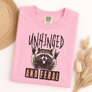 May include: A pink t-shirt with the words "UNHINGED" and "AND FERAL" printed in black. A raccoon graphic with its tongue out and hands in a rock and roll gesture is in the center of the shirt.