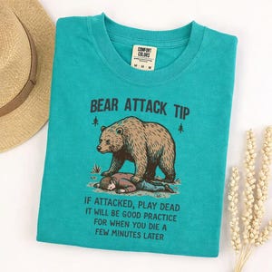 May include: Teal t-shirt with a humorous graphic. The design features a bear standing over a person, with the text "BEAR ATTACK TIP" above and the phrase "IF ATTACKED, PLAY DEAD..." below. The shirt has a crew neck and short sleeves.