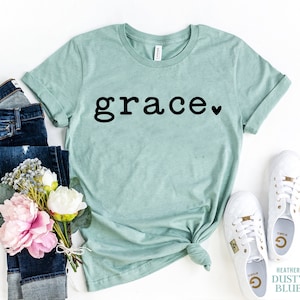 Pu&ograve; includere: Una t-shirt color blu polvere heather con la parola "grace" in testo nero e un piccolo cuore nero.