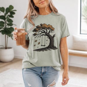 May include: A sage green short-sleeve t-shirt featuring a graphic illustration of a large tree with brown leaves and a swing hanging from a branch. The design includes several falling leaves and a dark shadow cast beneath the tree trunk.